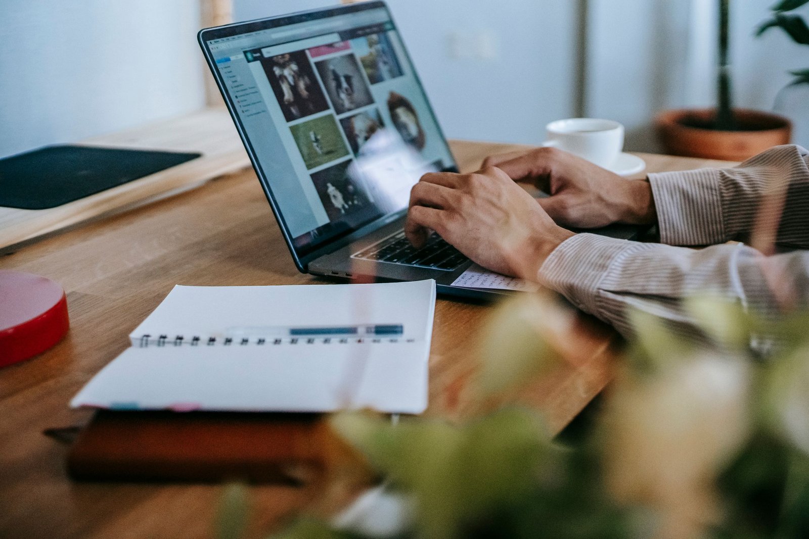 Crop unrecognizable guy in casual outfit working remotely on computer while sitting at wooden table near notebook with cup of coffee in light room